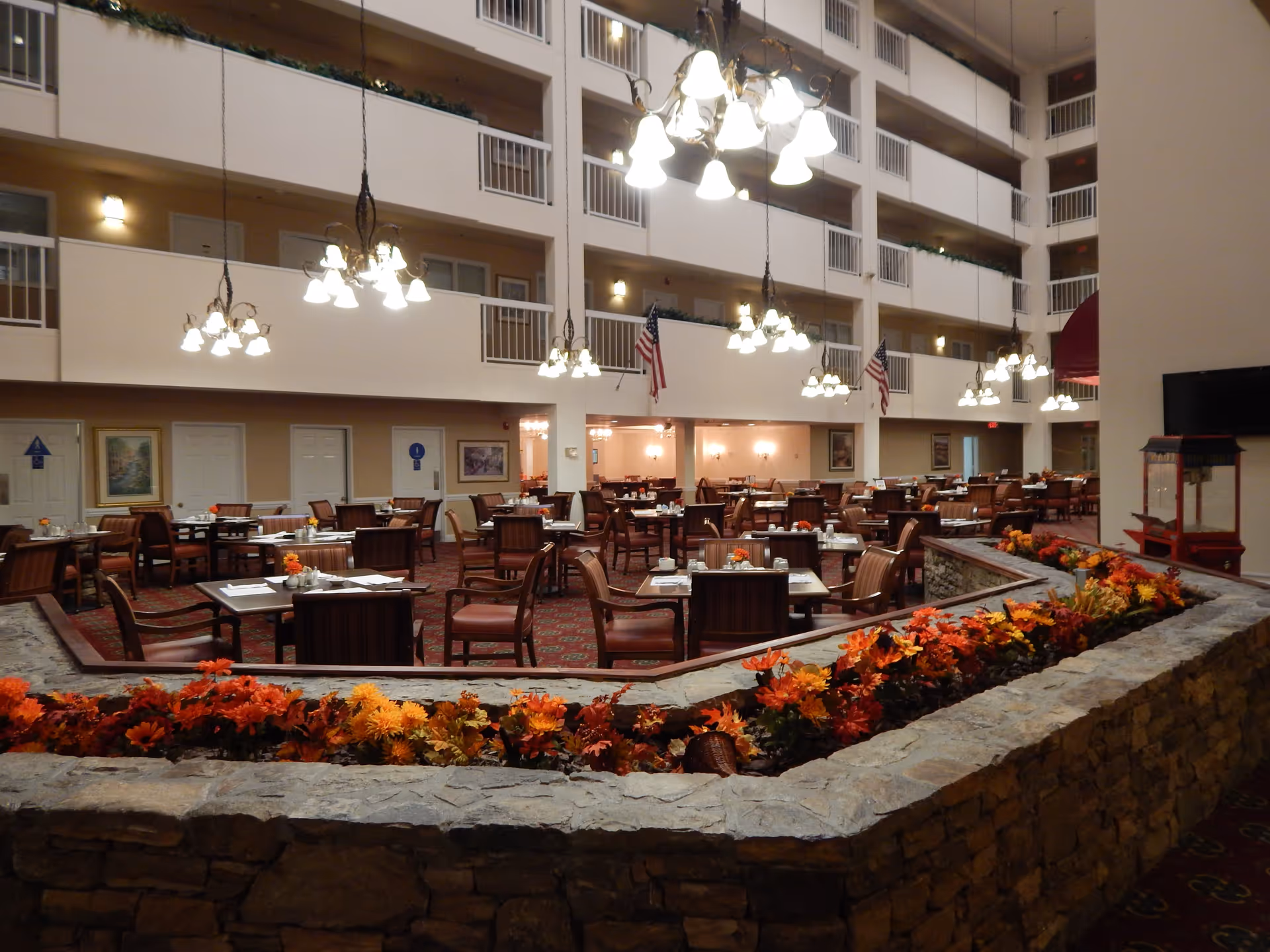 Interior view of a large dining area in a retirement community with multiple tables and chairs arranged neatly. The space features hanging chandeliers, American flags, and a stone planter filled with orange and yellow flowers in the foreground. The dining area is surrounded by multiple floors with railings and doors visible on the upper levels.