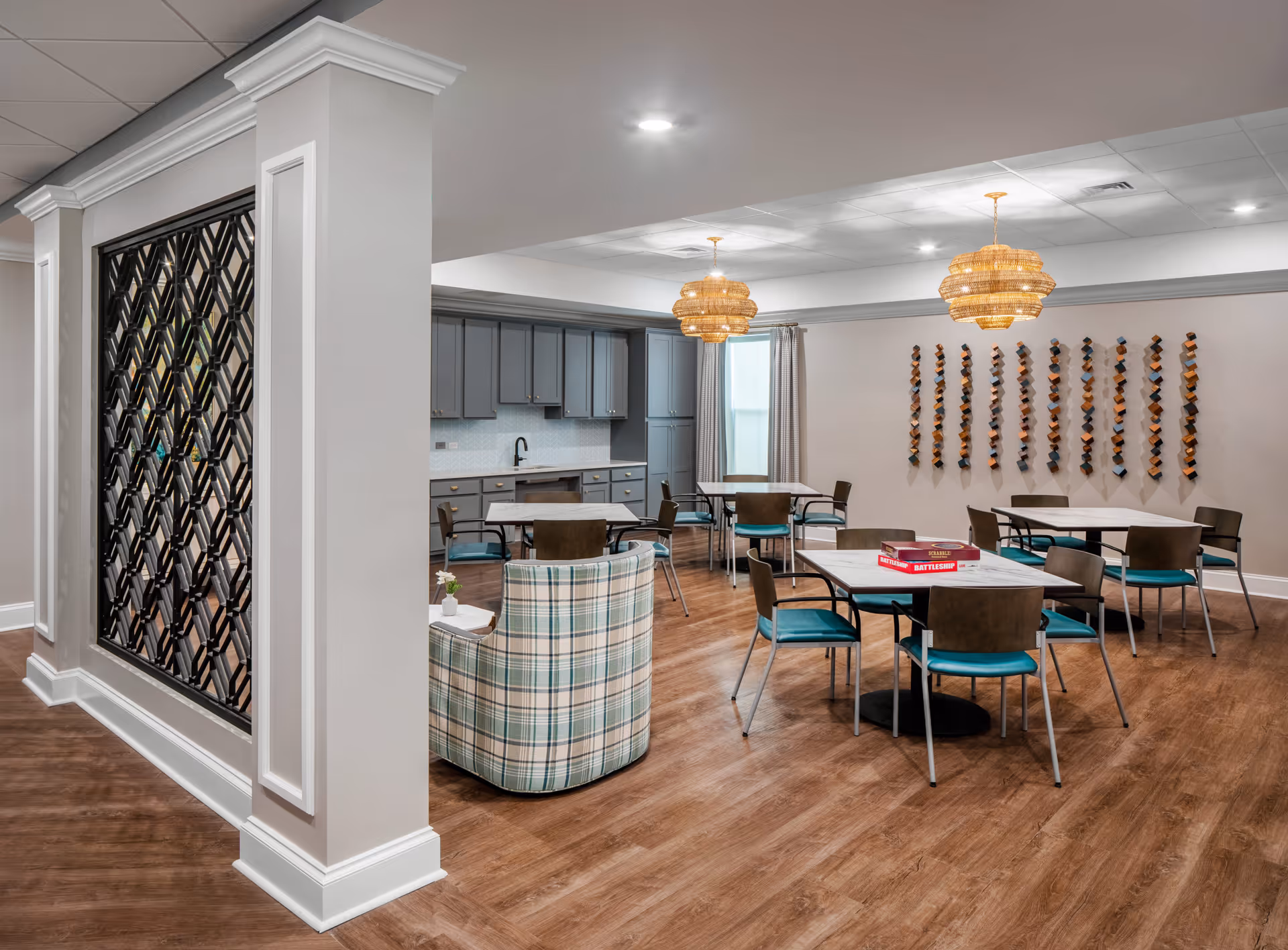 A bright and spacious common area with several tables and chairs arranged for group activities or dining. The room features a small kitchen area with gray cabinets and a sink. Two wicker pendant lights hang from the ceiling, and a decorative wine rack is mounted on the wall. A plaid upholstered armchair is positioned near the tables. The floor is wood, and the walls are painted light gray.