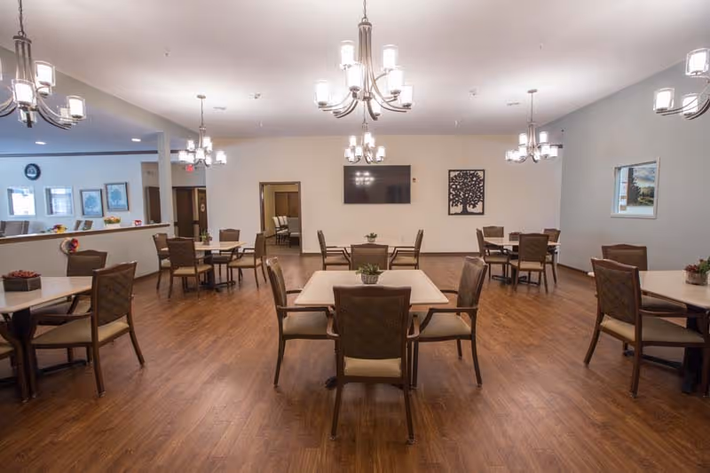 A spacious dining room with multiple square tables and cushioned chairs arranged neatly on a wooden floor. The room is well-lit with several modern chandeliers hanging from the ceiling. There is a flat-screen TV mounted on the far wall, along with a decorative wall art piece. The walls are painted in neutral tones, and there are windows and doorways visible in the background.