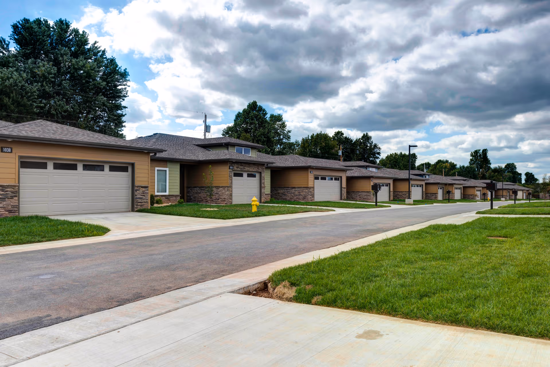 A row of single-story residential units with attached garages along a paved street, surrounded by green lawns and trees under a partly cloudy sky.