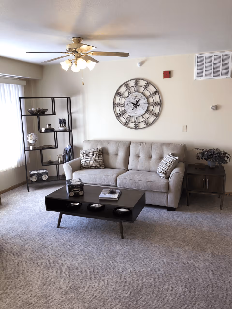 A cozy living room with a beige tufted sofa adorned with two patterned pillows, a dark wooden coffee table with decorative items, a matching side table with a plant, a black metal shelving unit with various decor pieces, a large round wall clock, and a ceiling fan with lights. The room has light-colored walls and carpeted flooring.