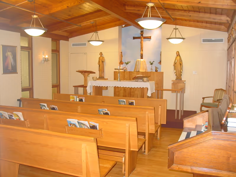 Interior of a small chapel with wooden pews, hymnals placed in the pew racks, a wooden altar covered with a white cloth, religious statues, a crucifix on the wall, and hanging ceiling lights under a wooden vaulted ceiling.