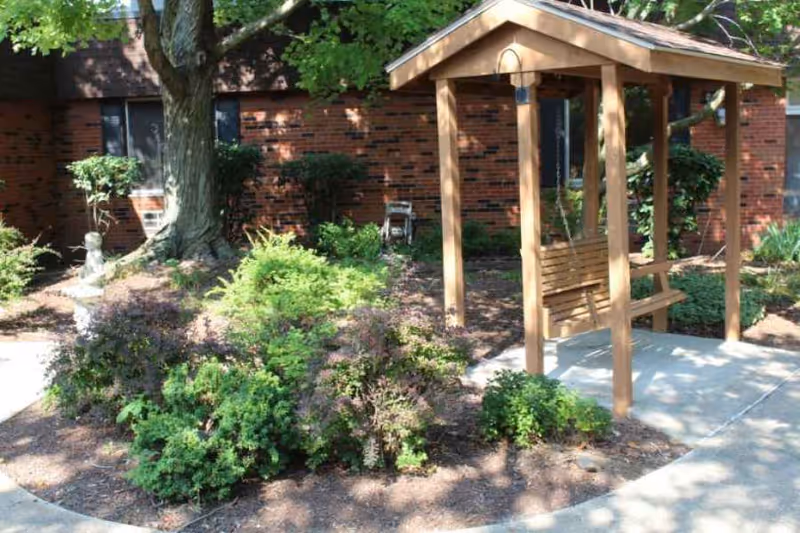 Outdoor garden area with a wooden swing under a small wooden pergola, surrounded by various green shrubs and plants. A large tree and a brick building with windows are visible in the background.