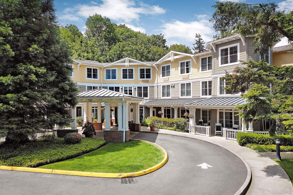 Exterior view of a senior living facility with a curved driveway leading to a covered entrance. The building is three stories tall with yellow and gray siding, white trim, and multiple windows. There are trees and well-maintained landscaping around the entrance area.