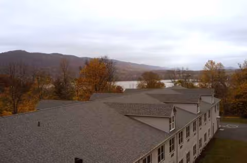 A multi-story senior living facility viewed from above, with autumn trees and a lake behind.
