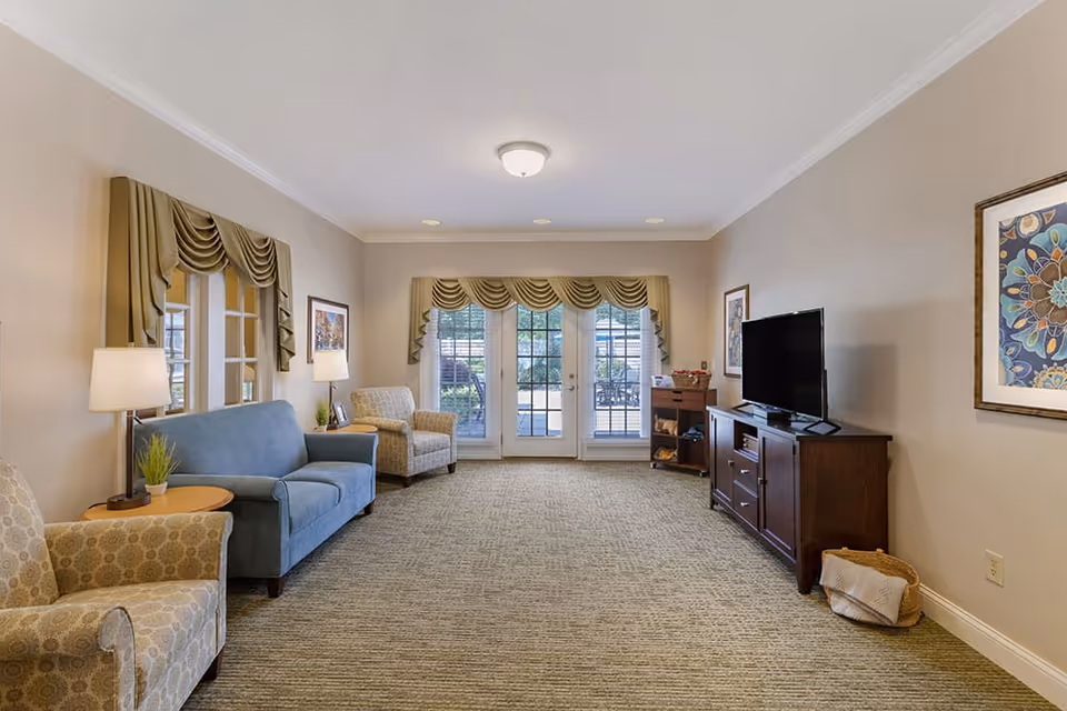 Bright furnished communal living room with sofas and armchairs, a TV on a wooden console, lamps, and French doors leading outside.