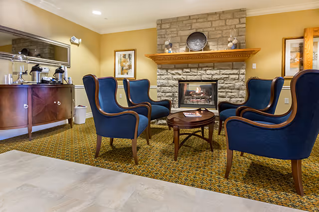 A cozy seating area with four blue upholstered armchairs arranged around a small wooden coffee table in front of a stone fireplace with a wooden mantel. The room has a patterned carpet, beige walls, framed artwork, and a wooden sideboard with coffee and tea supplies on the left side.