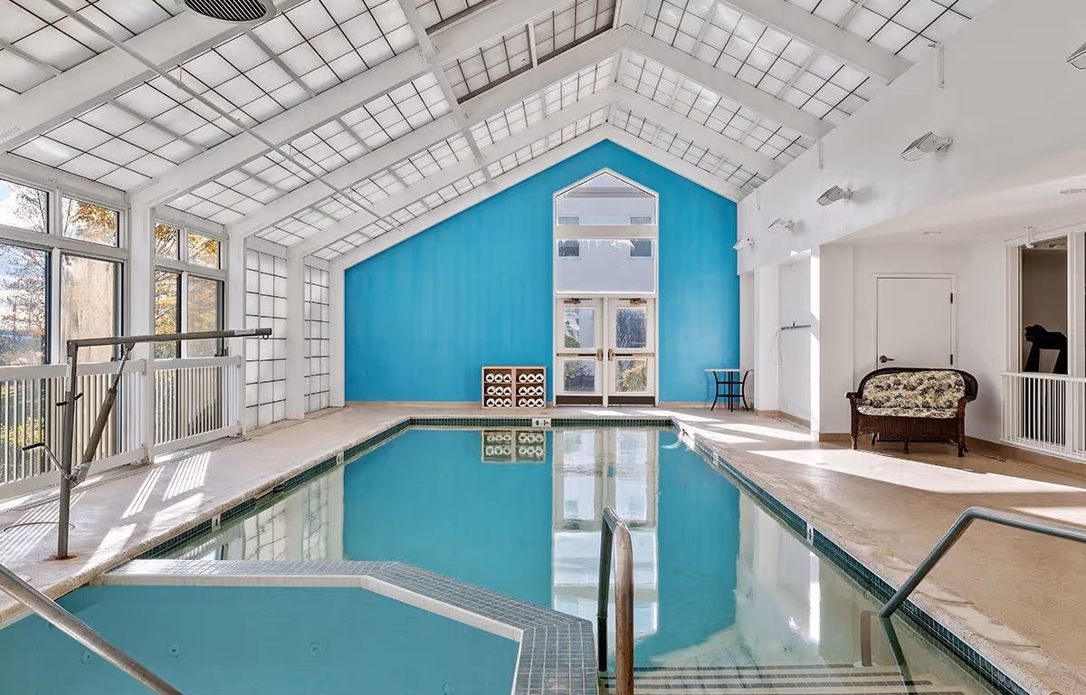 Bright indoor swimming pool with a vaulted glass ceiling, turquoise accent wall, seating and a pool lift.