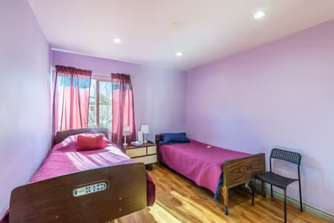 A small bedroom with two single beds, each with a wooden headboard and purple bedding. There is a nightstand with a lamp and a phone between the beds. A window with sheer purple curtains lets in natural light. A black chair is placed against the right wall. The walls are painted light purple and the floor is wooden.