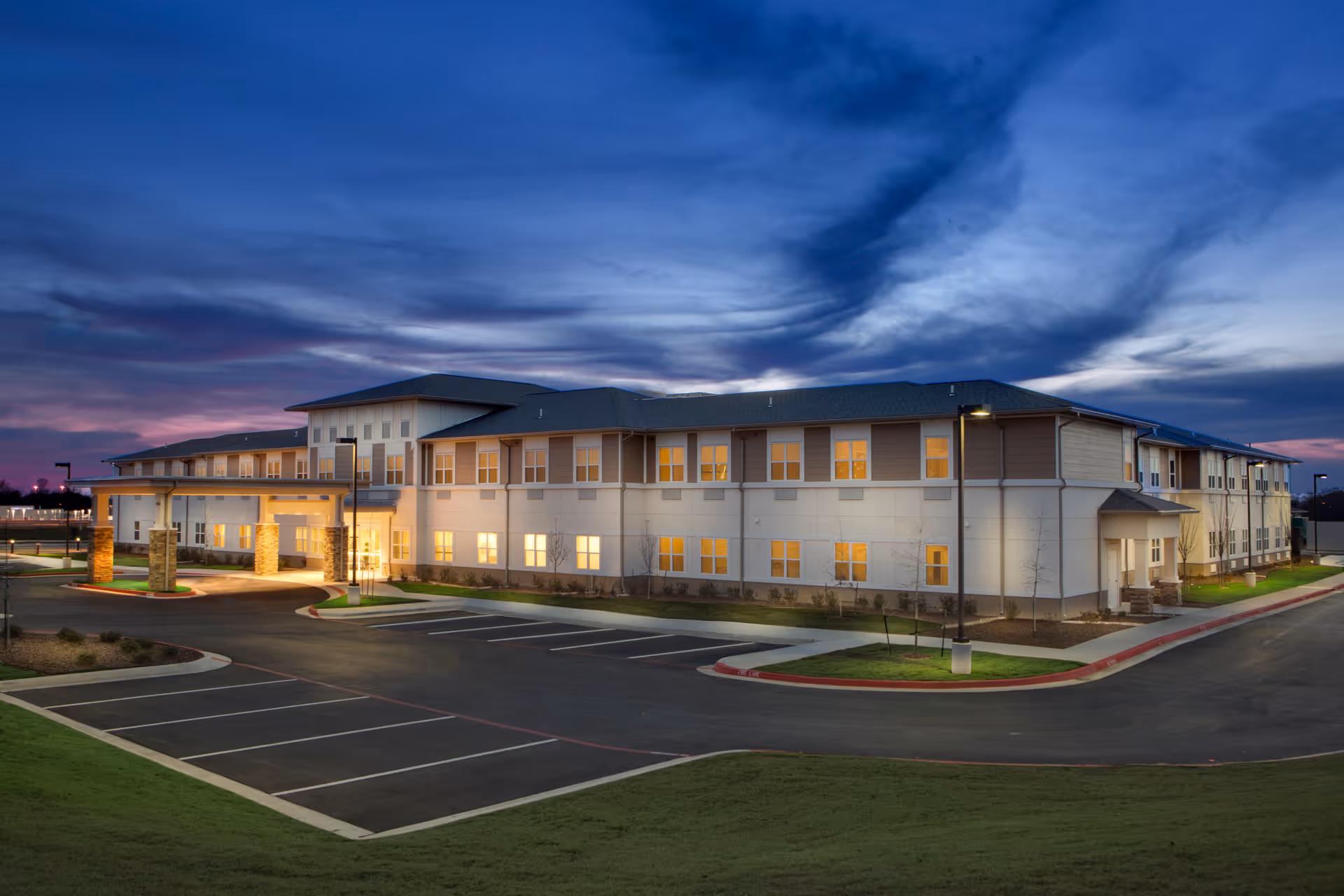 Exterior view of Prairie House senior living facility at dusk with lights glowing from windows, a large parking lot in front, and a dramatic cloudy sky.