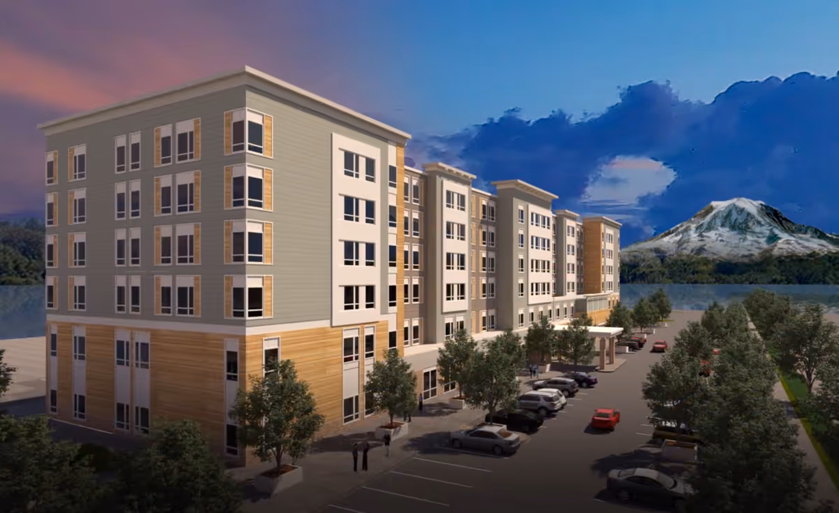 Exterior view of a multi-story senior living facility building named GenCare Lifestyle Tacoma at Point Ruston during dusk, with a parking lot in front, several parked cars, trees lining the walkway, and a mountain visible in the background under a partly cloudy sky.