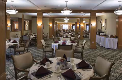 A spacious dining room with multiple round tables covered with white tablecloths, each set with plates, cups, silverware, and folded dark napkins. The room features beige walls, wooden columns, patterned carpet, and ceiling lights providing warm illumination. A buffet table with dishes and serving utensils is visible on the right side.