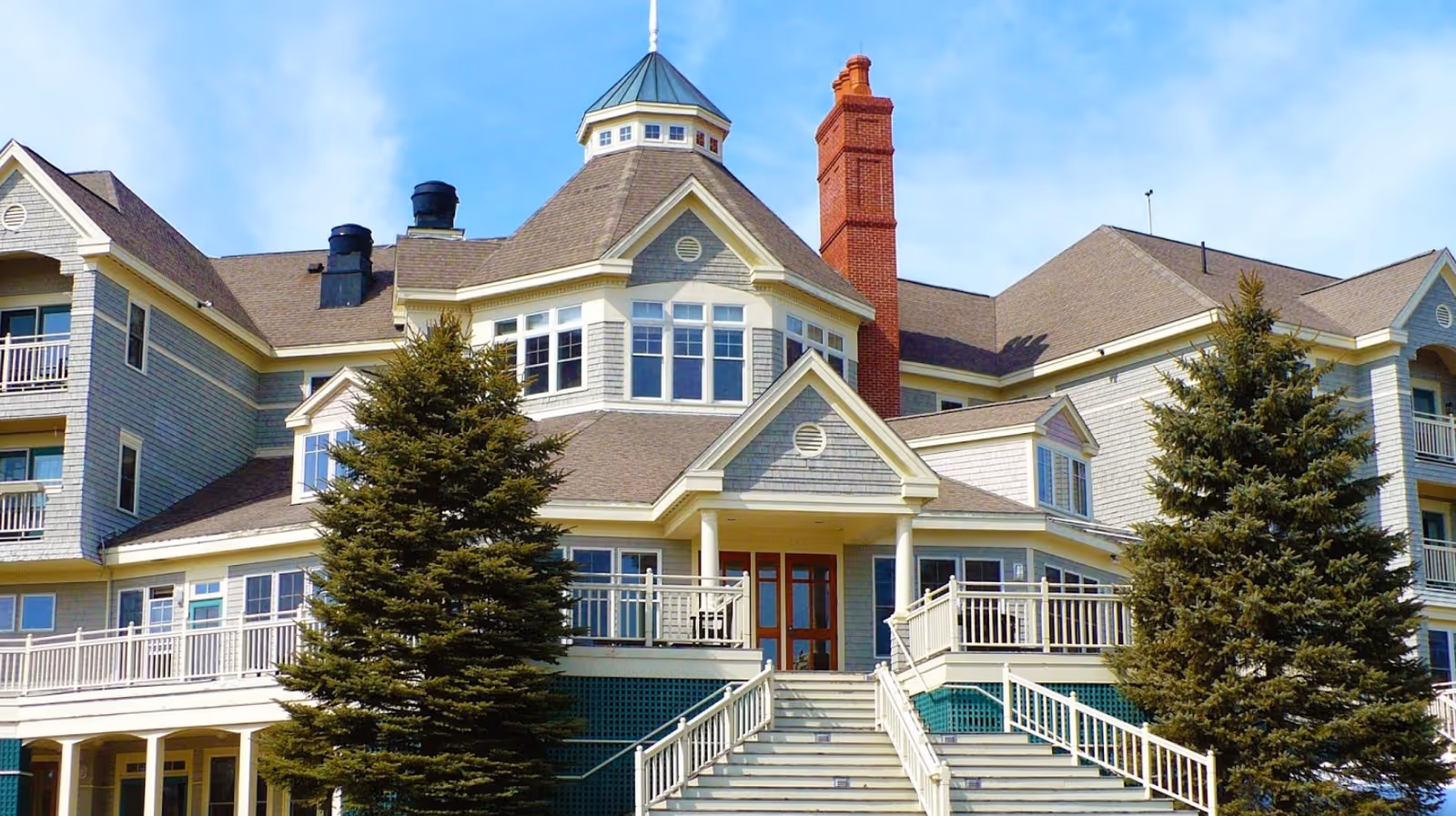 Front view of a large multi-story residential building with a central entrance, stairway, wraparound balconies, and evergreen trees.