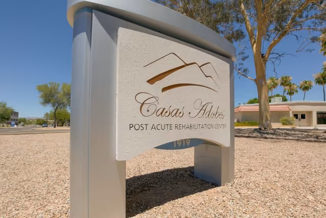 Outdoor sign for Casas Adobes Post Acute Rehabilitation Center with a stylized mountain logo, set in a gravel area with trees and a building in the background under a clear blue sky.
