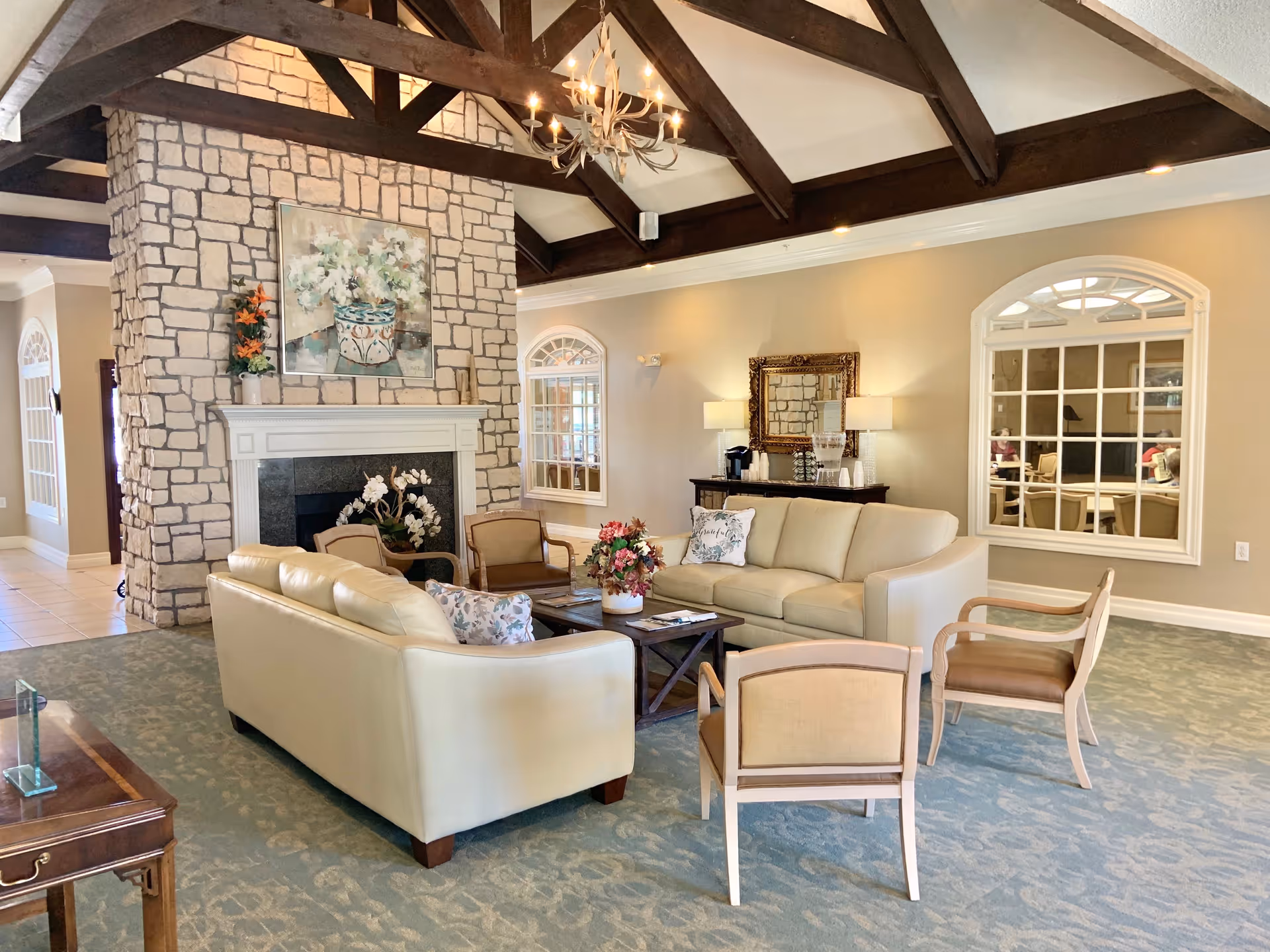 A spacious and well-lit living room area with beige sofas and chairs arranged around a wooden coffee table. The room features a large stone fireplace with a floral painting above it, exposed dark wooden ceiling beams, a chandelier, and large windows with white trim. There is a sideboard with a mirror and lamps against the wall, and the carpet has a subtle pattern.