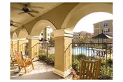 Covered arched patio with rocking chairs and ceiling fans overlooking a swimming pool and surrounding buildings.