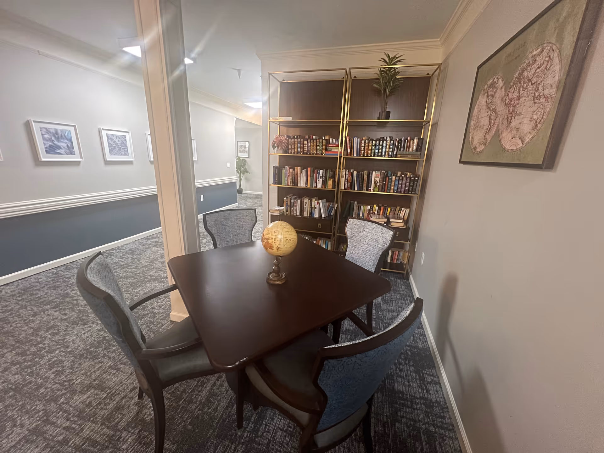 A small reading or meeting area in a senior living facility with a square wooden table surrounded by four upholstered chairs. A decorative globe sits on the table. Behind the table is a tall bookshelf filled with books and a small potted plant on top. The walls are decorated with framed pictures and a vintage-style world map. The carpet is gray with a textured pattern, and the hallway outside the room is visible with additional framed artwork on the walls.