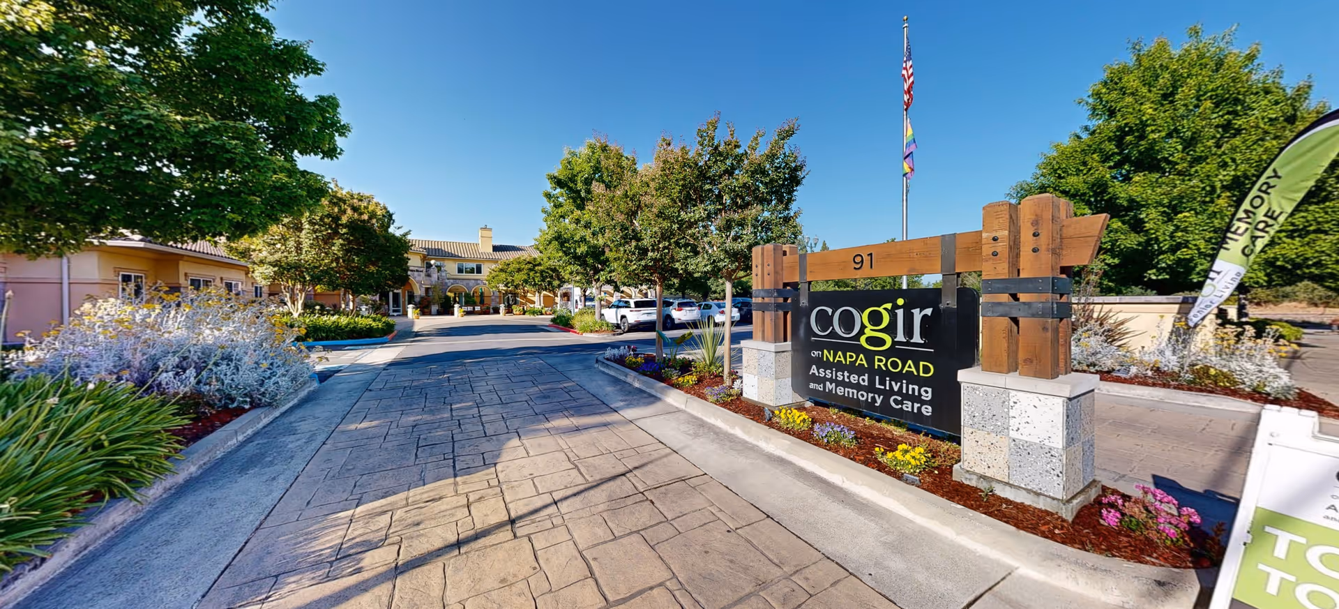 Entrance driveway to Cogir of Sonoma Plaza assisted living and memory care facility with a large wooden sign displaying the facility name, surrounded by landscaped plants and trees under a clear blue sky.