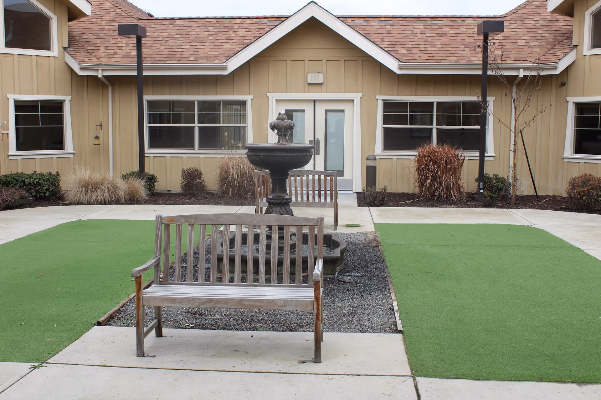 Courtyard with wooden benches and a central fountain in front of a beige single-story building.
