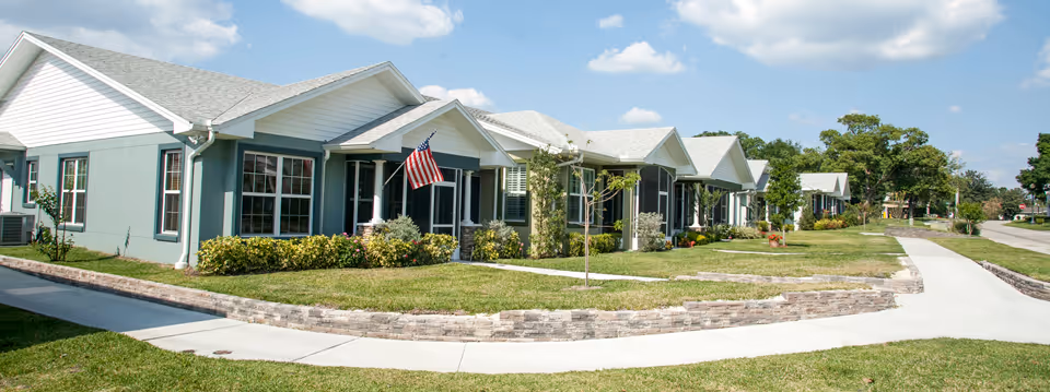 Row of single-story residential buildings with light blue and white exteriors, manicured lawns, small trees, and an American flag displayed on one porch under a partly cloudy sky.