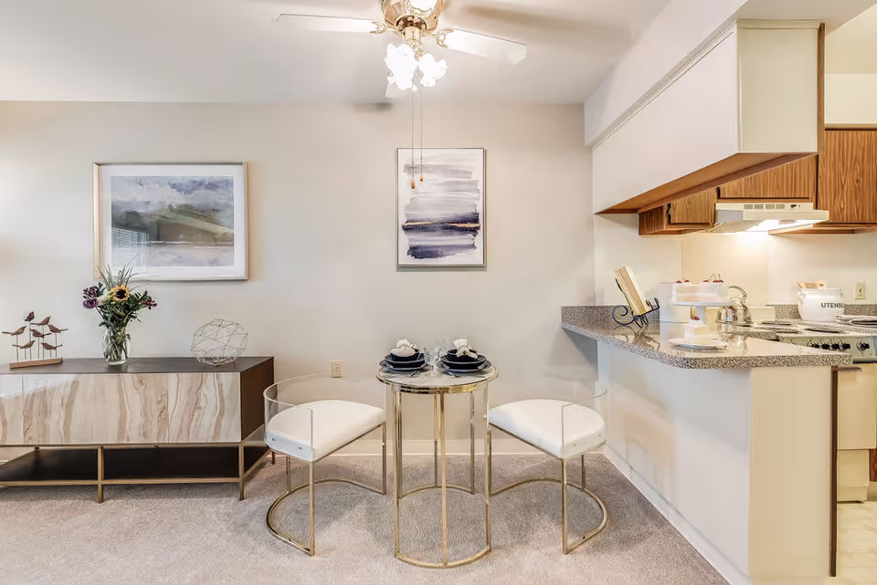 A small dining area with a round glass table set for two with black plates and white napkins. Two white cushioned chairs with gold metal frames are placed around the table. To the left, there is a wooden sideboard with decorative items including a vase with flowers, a metal geometric sculpture, and a small bird sculpture. The wall behind has two framed abstract paintings. To the right, a kitchen counter with a granite countertop extends into the dining area, with a stove and kitchen utensils visible in the background. The room is carpeted and well-lit with a ceiling fan overhead.