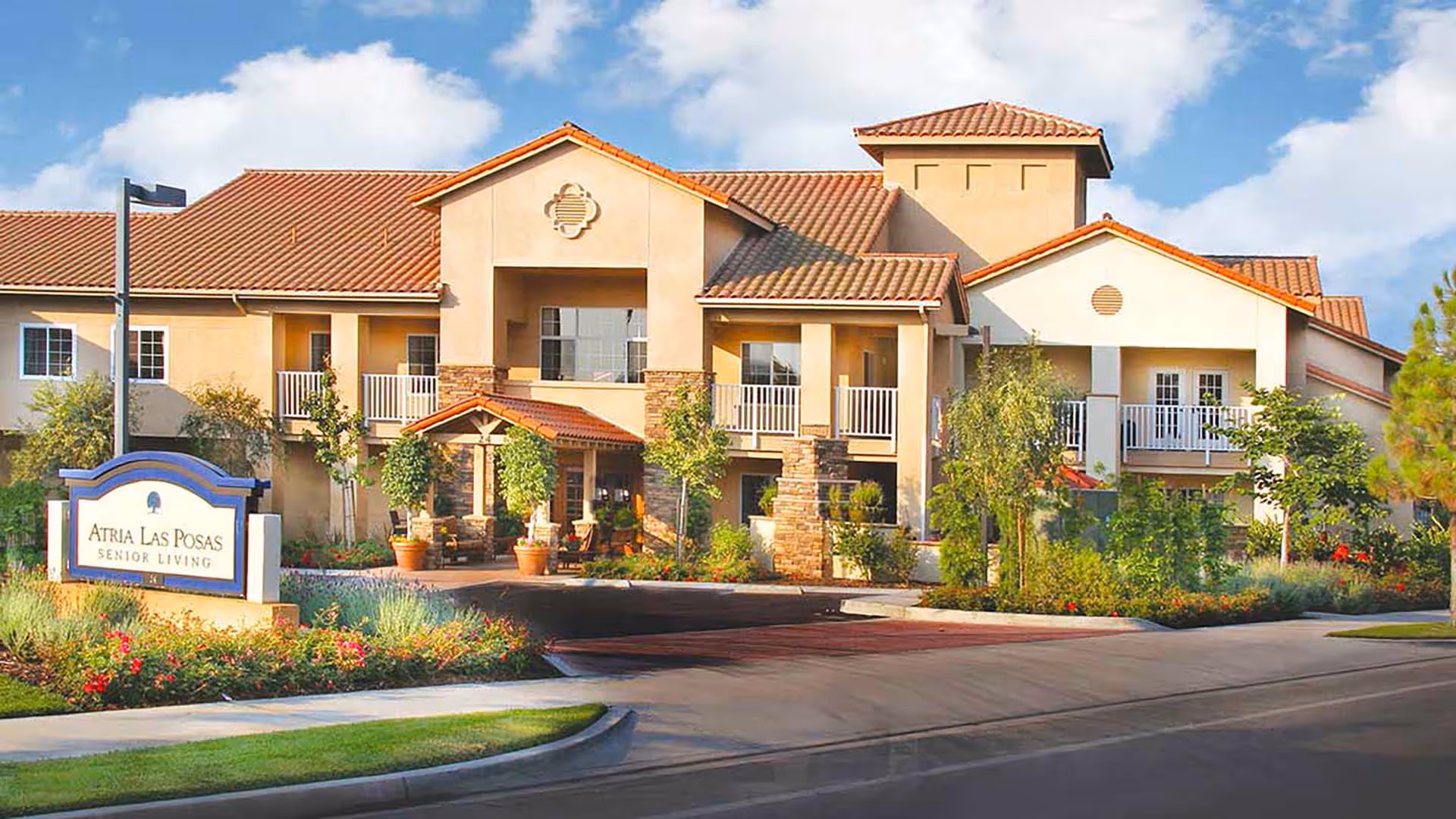 Exterior view of Atria Las Posas senior living facility showing a two-story building with balconies, a tiled roof, landscaped garden with flowers and trees, and a sign in front that reads 'Atria Las Posas Senior Living'.