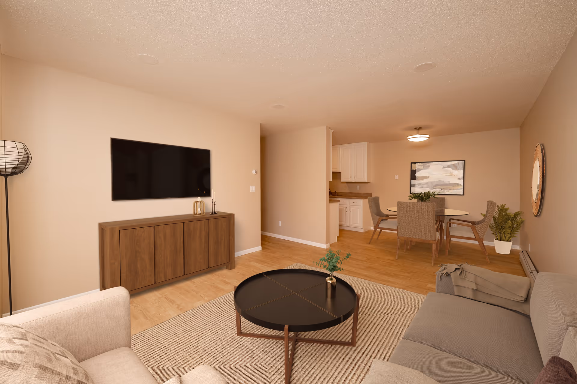 A cozy living room and dining area in a senior living facility. The living room features a beige sofa with a throw blanket, a round black coffee table with a small plant, a wooden cabinet with decorative items, and a wall-mounted flat-screen TV. The dining area has a round table with four upholstered chairs, a framed abstract painting on the wall, and a potted plant in the corner. The space has light wood flooring and neutral-colored walls.