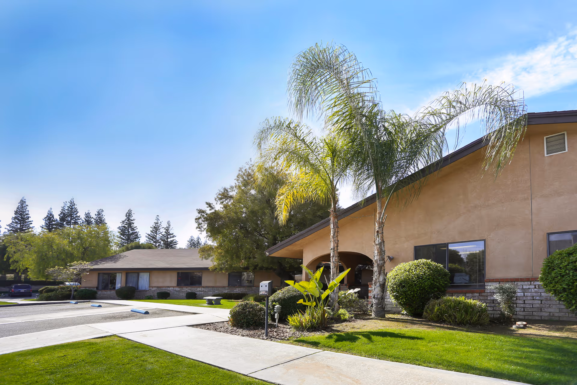 Exterior view of Bakersfield Post Acute facility showing a single-story building with beige walls, several windows, and a sloped roof. The foreground features a well-maintained lawn, palm trees, bushes, and a paved walkway under a clear blue sky.