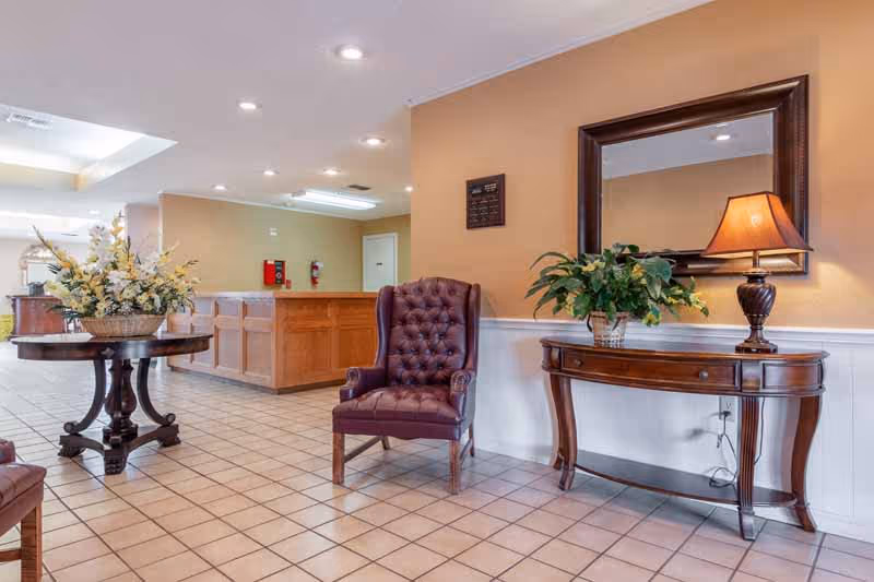 A well-lit interior space with tiled flooring featuring a wooden reception desk in the background, a round wooden table with a floral arrangement, a dark leather tufted armchair, and a wooden console table with a lamp and a potted plant beneath a large framed mirror on a beige wall.
