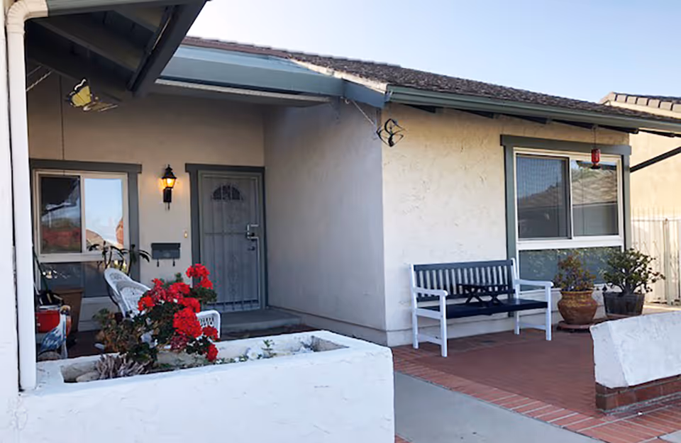 Front porch of a single-story home with a bench, potted plants, red flowers and a screened front door.