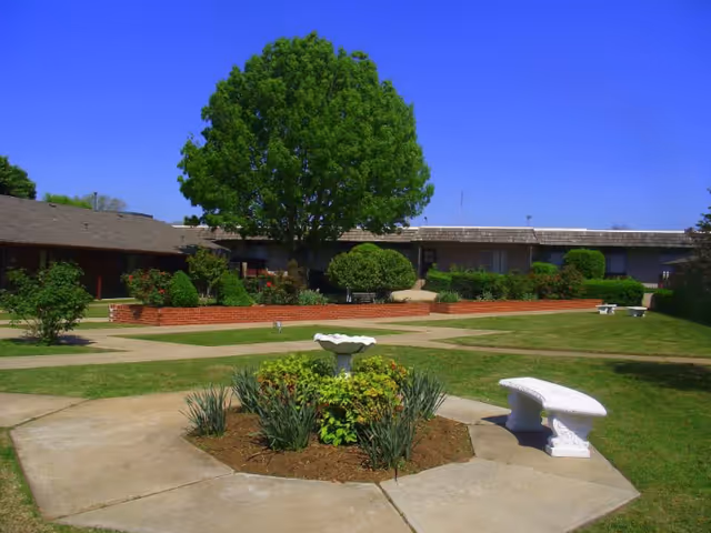 Outdoor courtyard area with a large green tree, trimmed bushes, and a circular flower bed with a birdbath in the center. There are concrete walkways and white stone benches around the garden area, with a single-story building in the background under a clear blue sky.