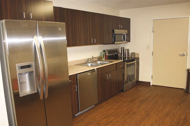 Modern kitchen area with stainless steel refrigerator, dishwasher, oven, and microwave. The kitchen has dark wood cabinets, a beige countertop, and a wooden floor. There is a closed door at the end of the kitchen space.