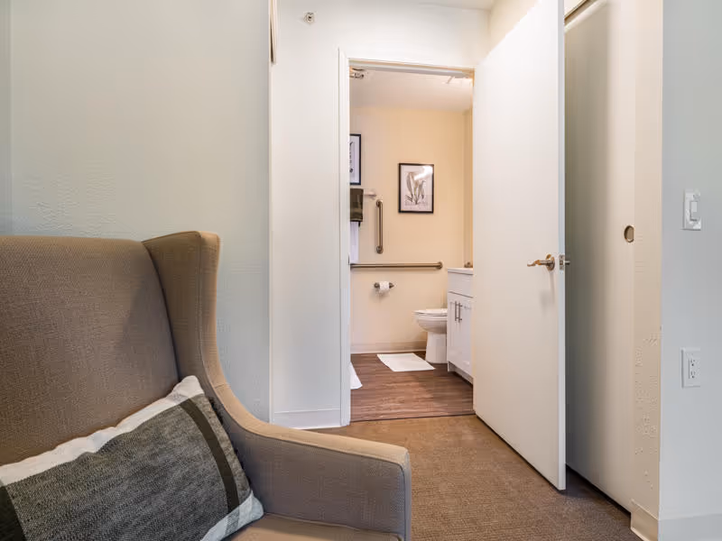 View of a beige armchair with a cushion in the foreground, positioned near an open door leading to a bathroom. The bathroom features a toilet, white vanity with a sink, a towel rack with a green towel, two framed botanical prints on the wall, and a wooden floor with white bath mats.