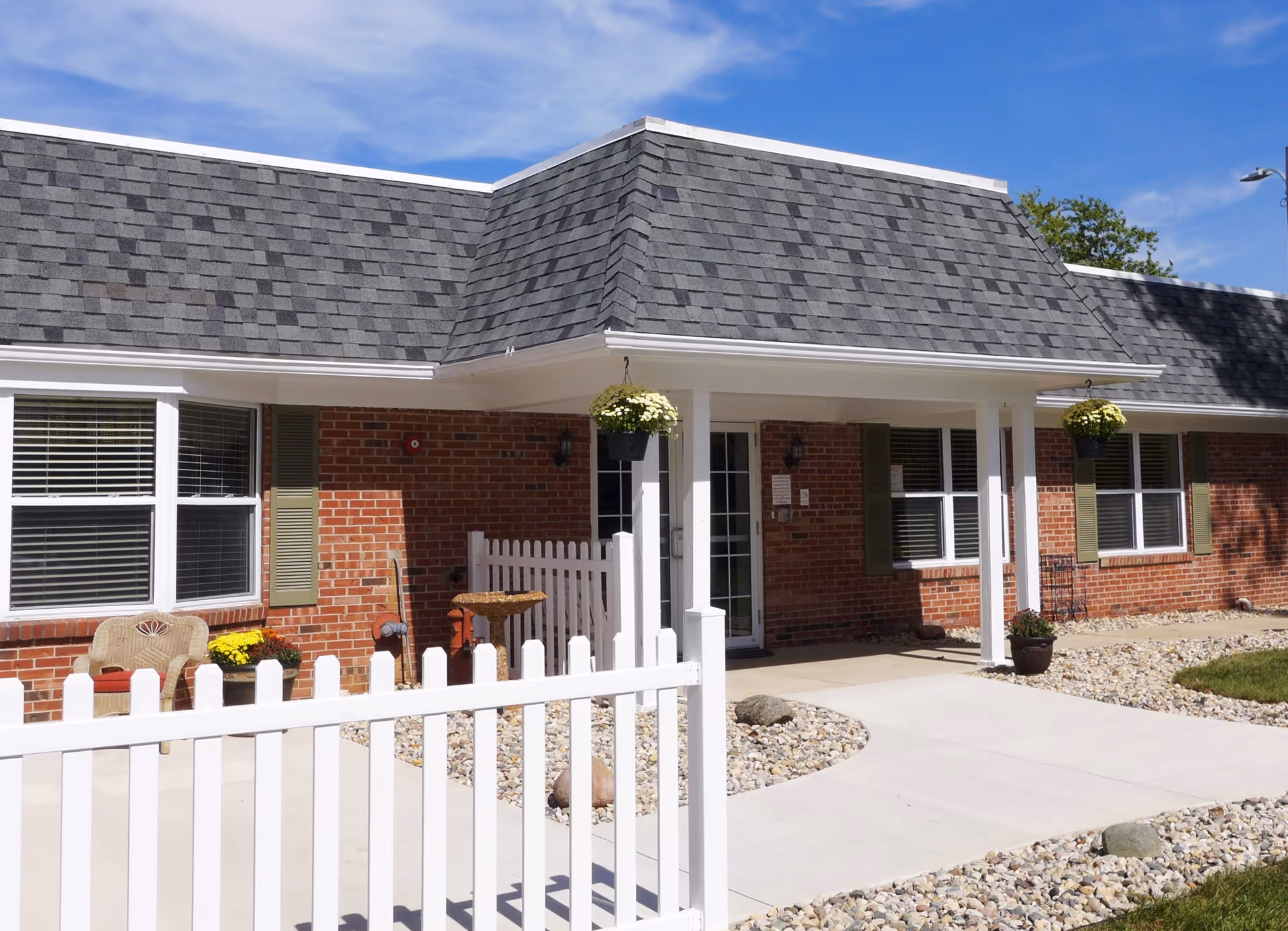 Front entrance of a single-story brick senior living building with a white picket fence, covered porch, and hanging planters.