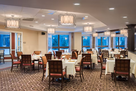 A spacious dining room with multiple tables covered in white tablecloths, each set with glasses, plates, and silverware. The room features patterned carpet flooring, large windows letting in natural light, and modern pendant lights hanging from the ceiling. Chairs with cushioned seats and backs surround each table.