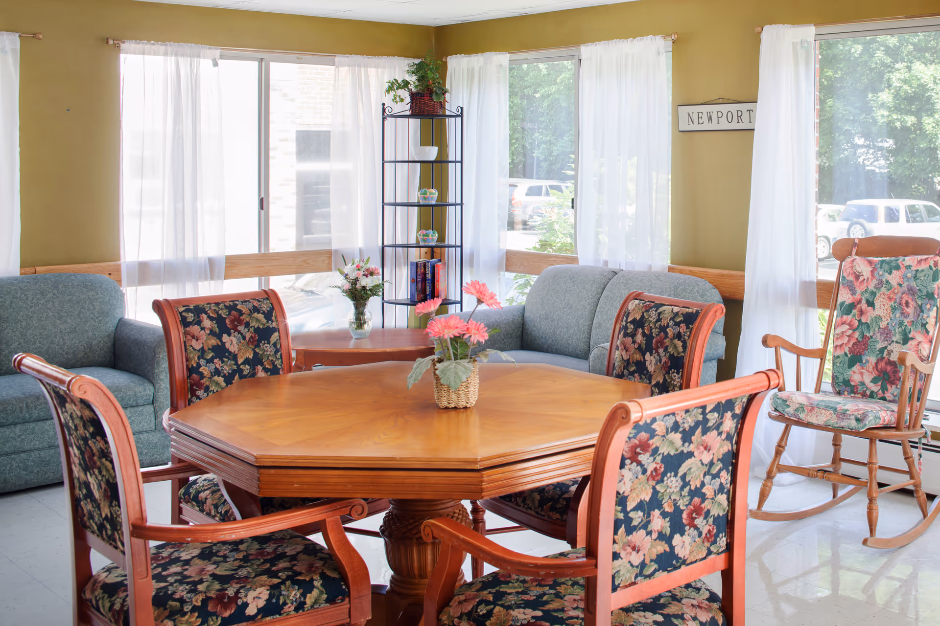A bright living room with large windows covered by sheer white curtains. The room features a wooden hexagonal table surrounded by four floral upholstered chairs, a floral rocking chair, and two green upholstered sofas. A small black metal shelf with decorative items and books is placed in the corner. A small flower arrangement is on the table, and a sign reading 'NEWPORT' is on the wall.