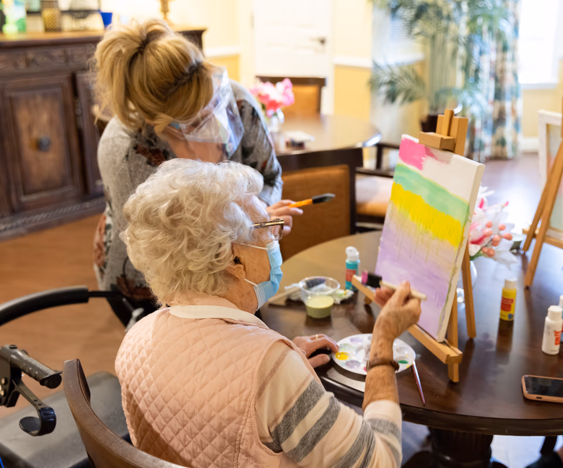 An elderly woman wearing a face mask is painting on a canvas set on an easel at a table. A caregiver or instructor wearing a face shield is standing beside her, assisting with the painting activity. The room has warm lighting and furniture including a wooden cabinet and a round table with art supplies.