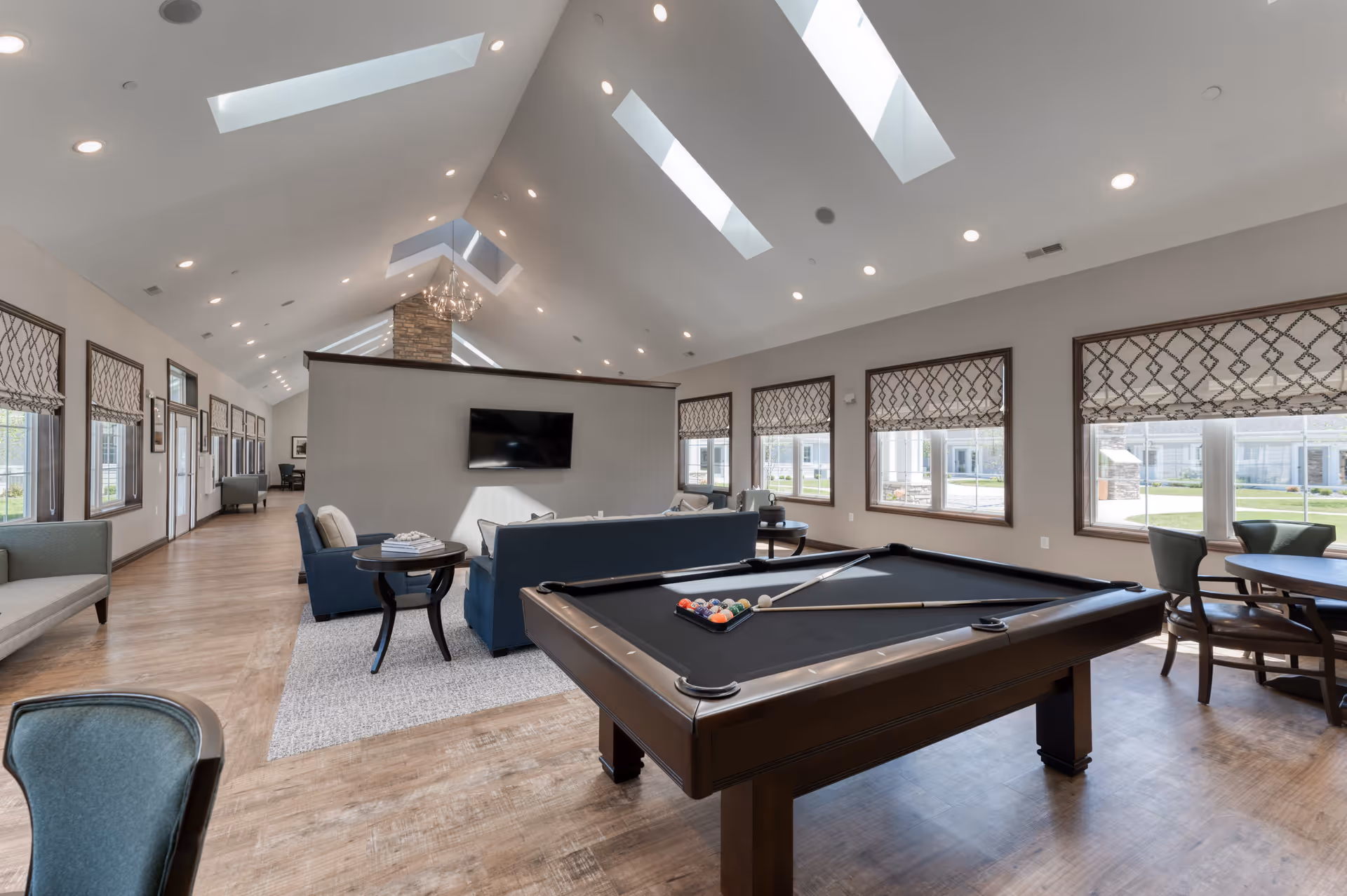 Bright communal lounge with a pool table, seating area, wall-mounted TV, and large windows under skylights.