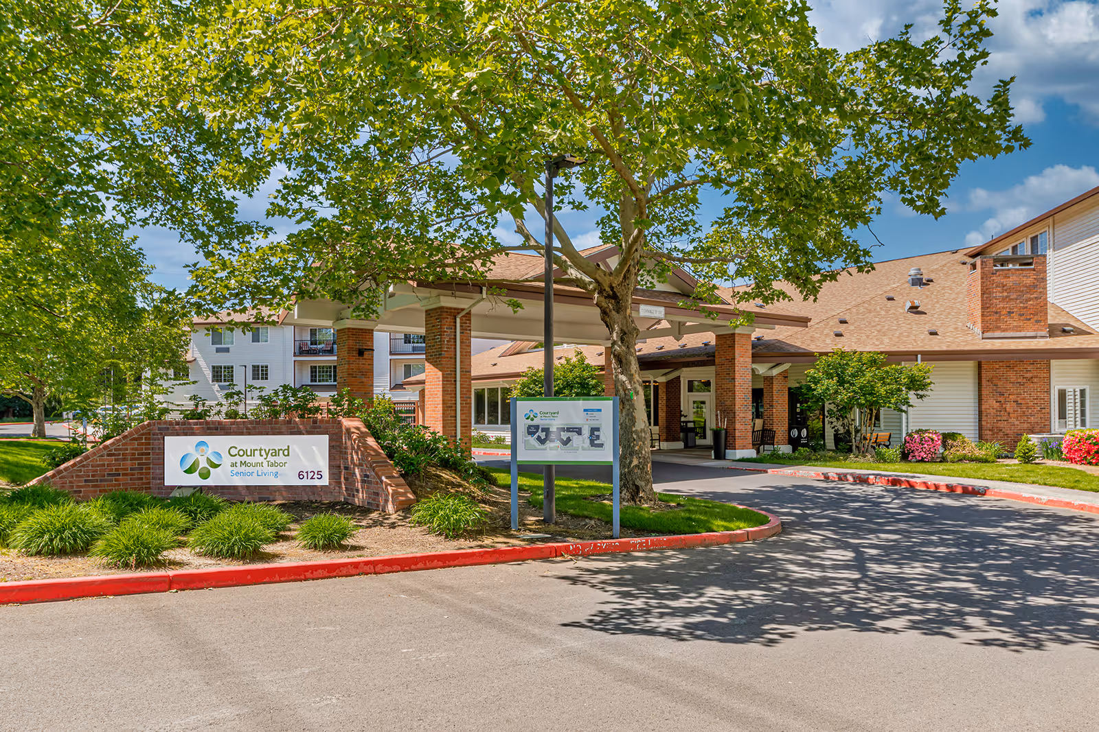 Exterior view of Courtyard at Mount Tabor senior living facility showing the entrance with a covered drop-off area, a large tree, landscaped greenery, and a sign with the facility name and address number 6125.