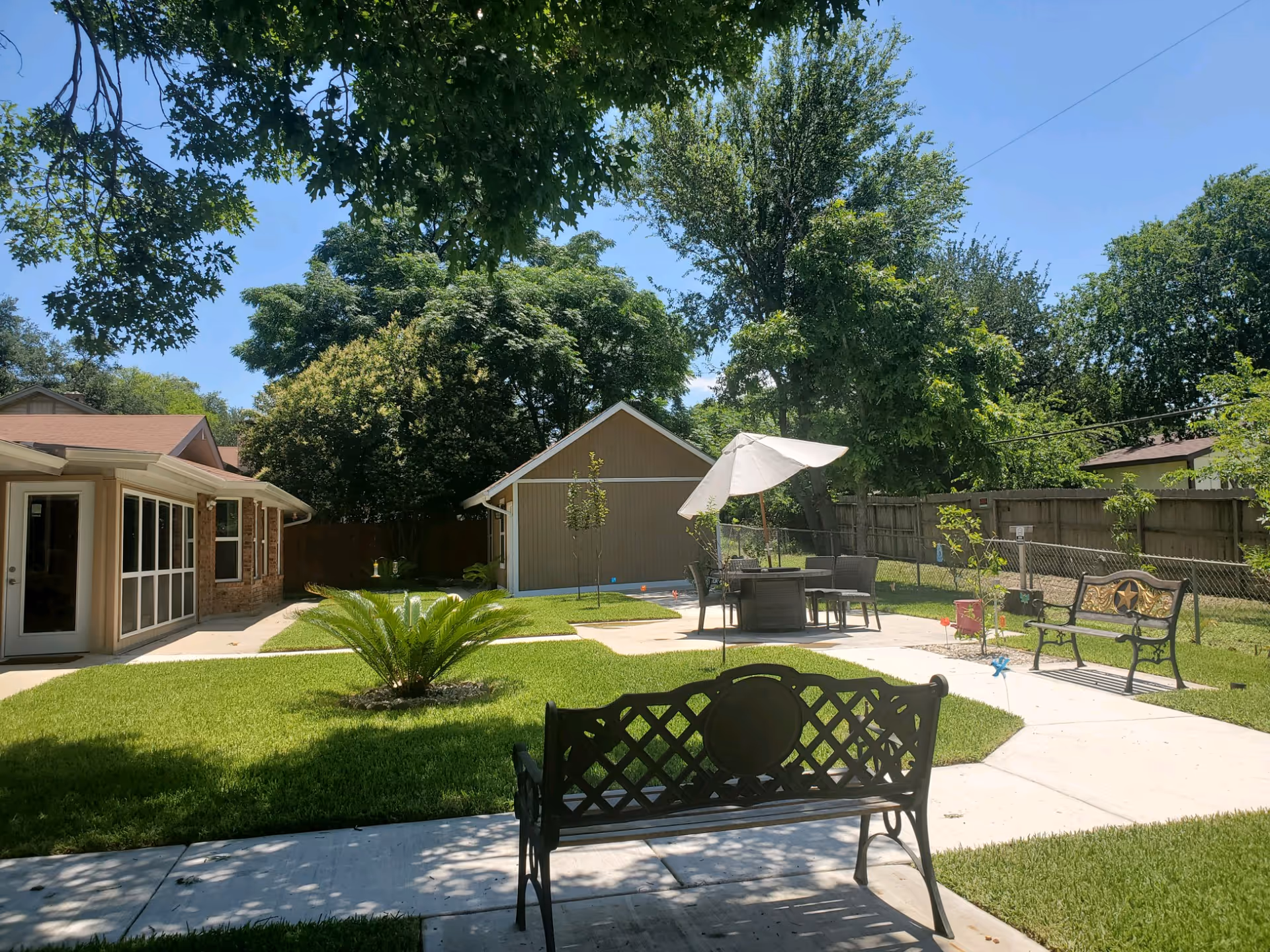 Well-maintained outdoor patio and lawn with benches, a table and umbrella, and a small outbuilding surrounded by trees.