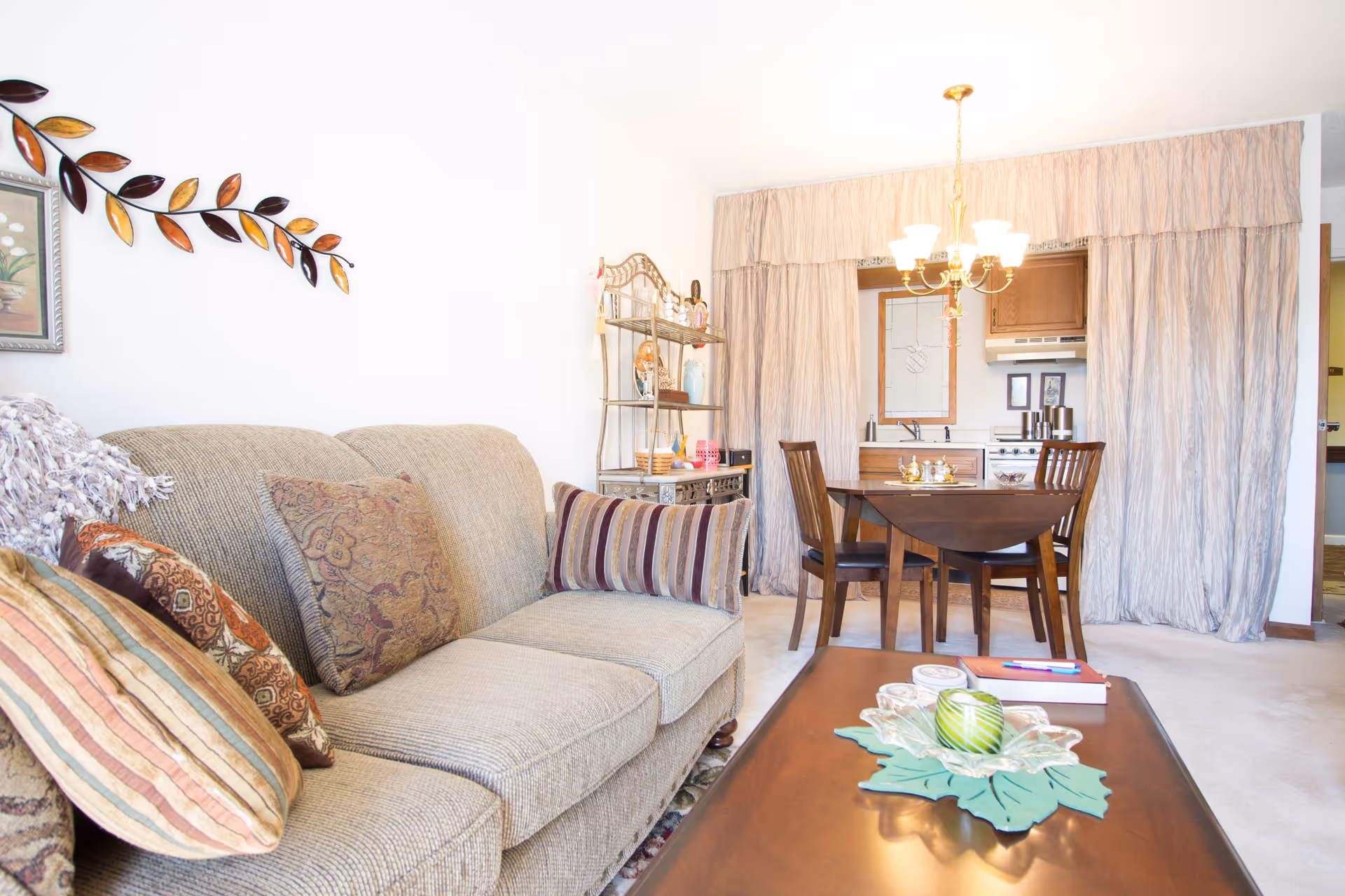 Bright living room with a beige sofa, coffee table, and a dining table with chairs in front of a curtained kitchenette.