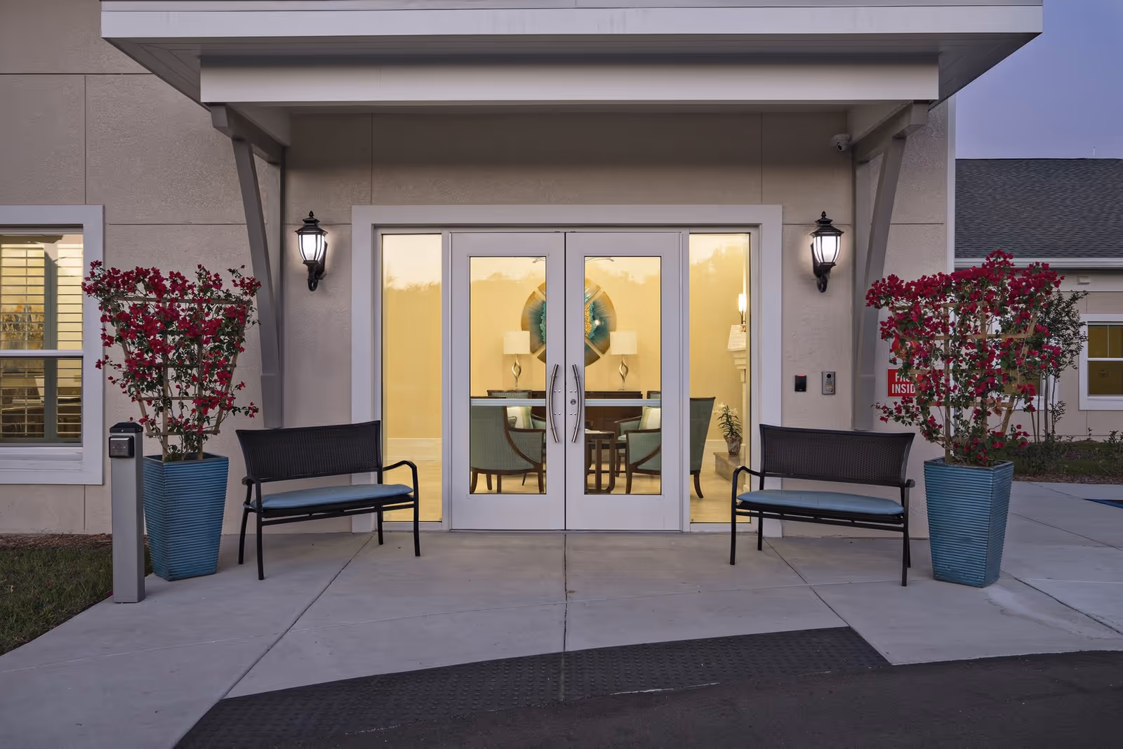Entrance to a building with double glass doors flanked by two black benches with blue cushions and two large blue planters containing red flowering plants. Two wall-mounted lantern-style lights are on either side of the doors. Inside, a room with chairs, a table, lamps, and wall decor is visible through the glass doors.