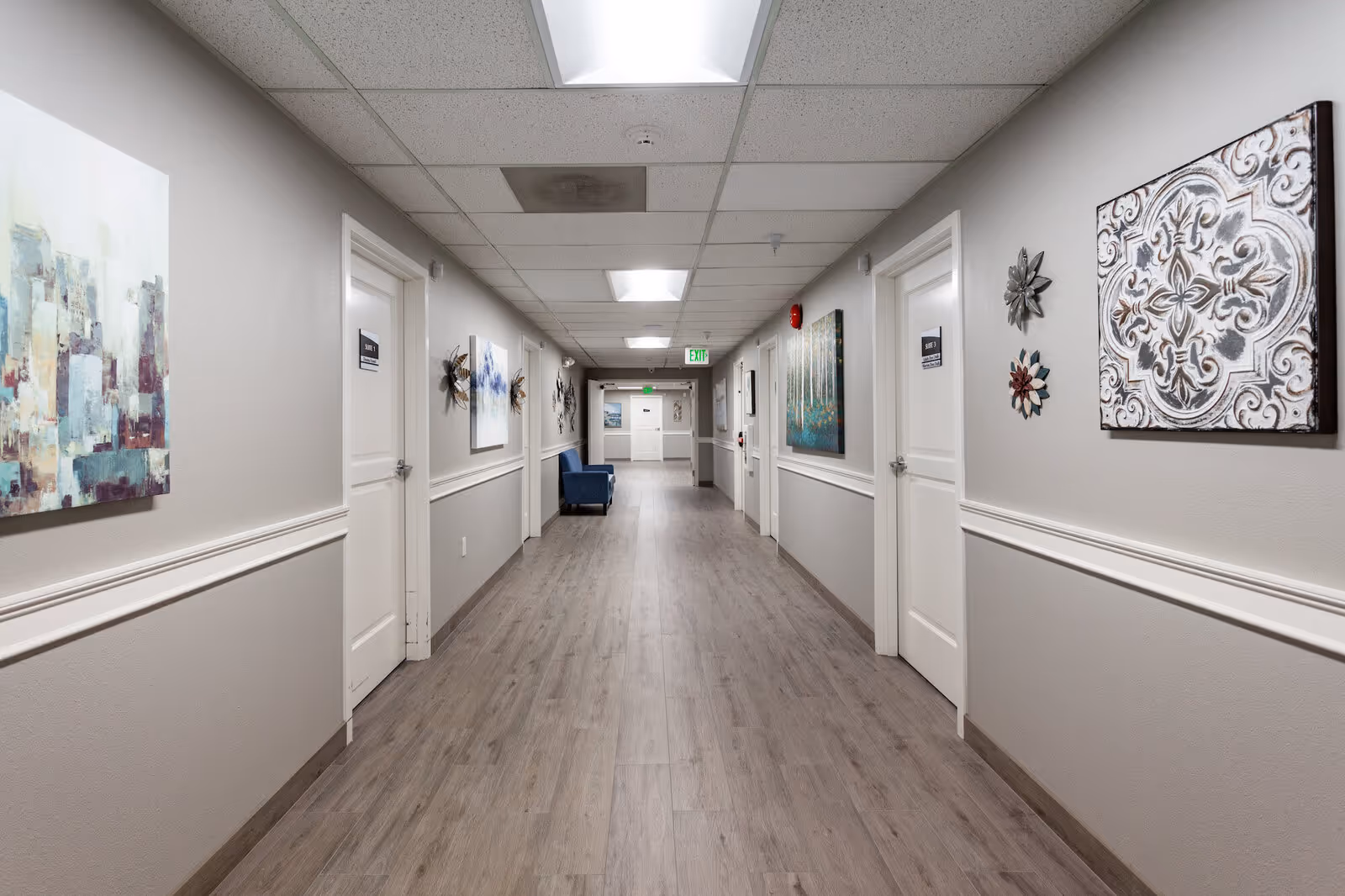 A long, well-lit interior hallway in an assisted living facility lined with doors, artwork, and seating.