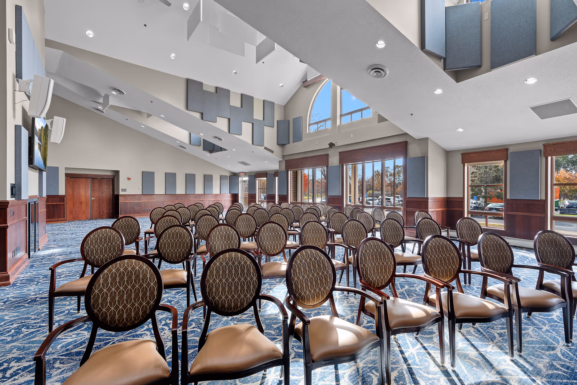 A spacious meeting or conference room with rows of patterned cushioned chairs arranged facing a wall-mounted television. The room features high ceilings with recessed lighting, large windows allowing natural light to enter, and blue and beige patterned carpet. The walls have wooden paneling and acoustic panels, and there is a large arched window near the ceiling.
