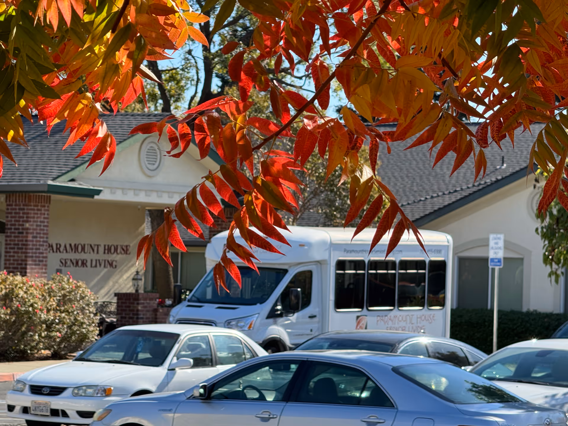 View of the exterior of Paramount House Senior Living facility with a white shuttle bus and several parked cars in front. Red and orange autumn leaves partially frame the top of the image.