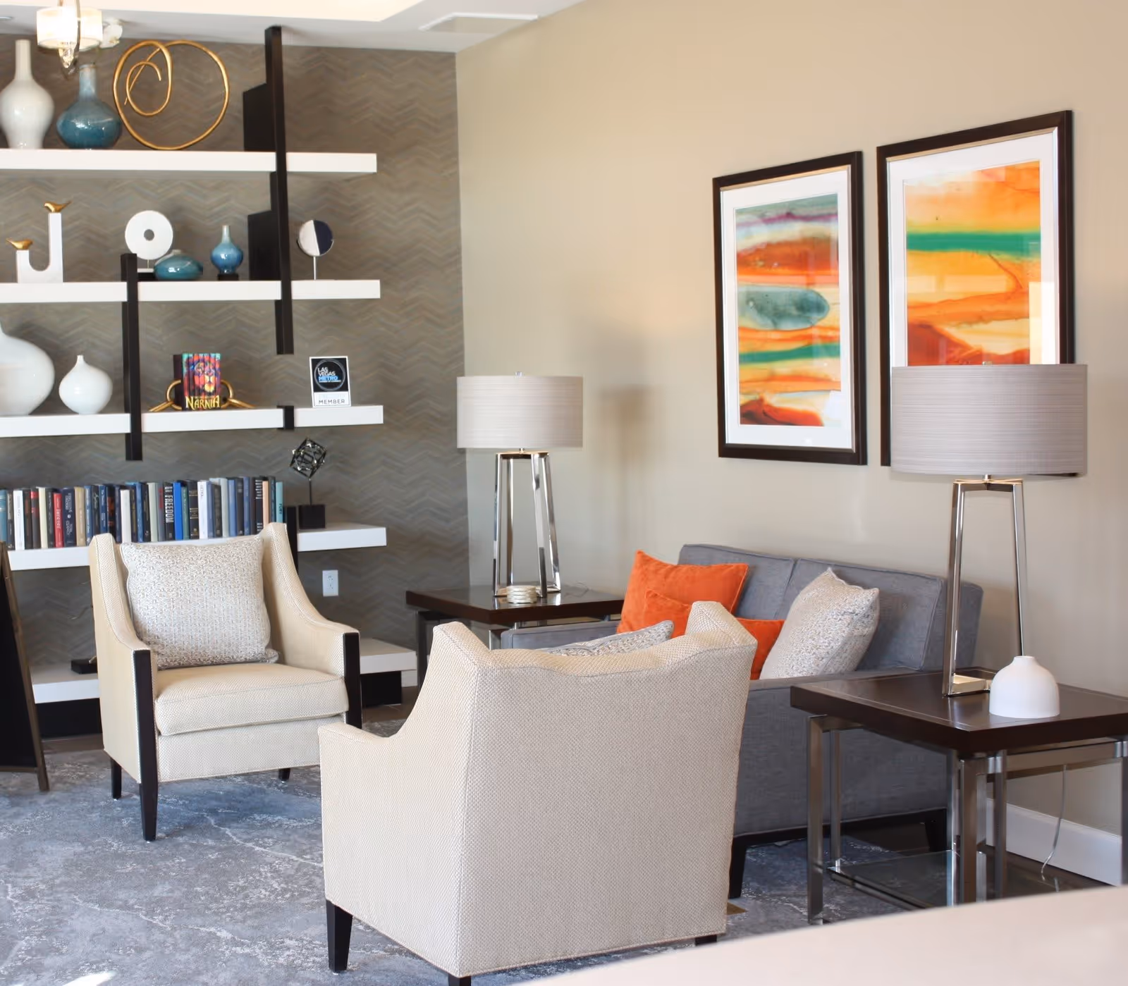 A cozy living room area with two beige armchairs and a gray sofa adorned with orange and beige pillows. There are two modern table lamps on dark wooden side tables, and two colorful abstract paintings hang on the beige wall. A decorative shelving unit with books, vases, and ornaments is visible in the background.