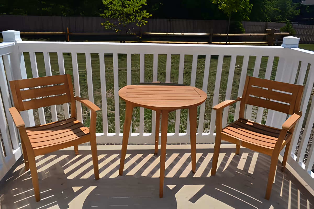 A small outdoor balcony or patio area with two wooden chairs and a matching round wooden table between them, surrounded by a white railing. In the background, there is green grass and some trees.