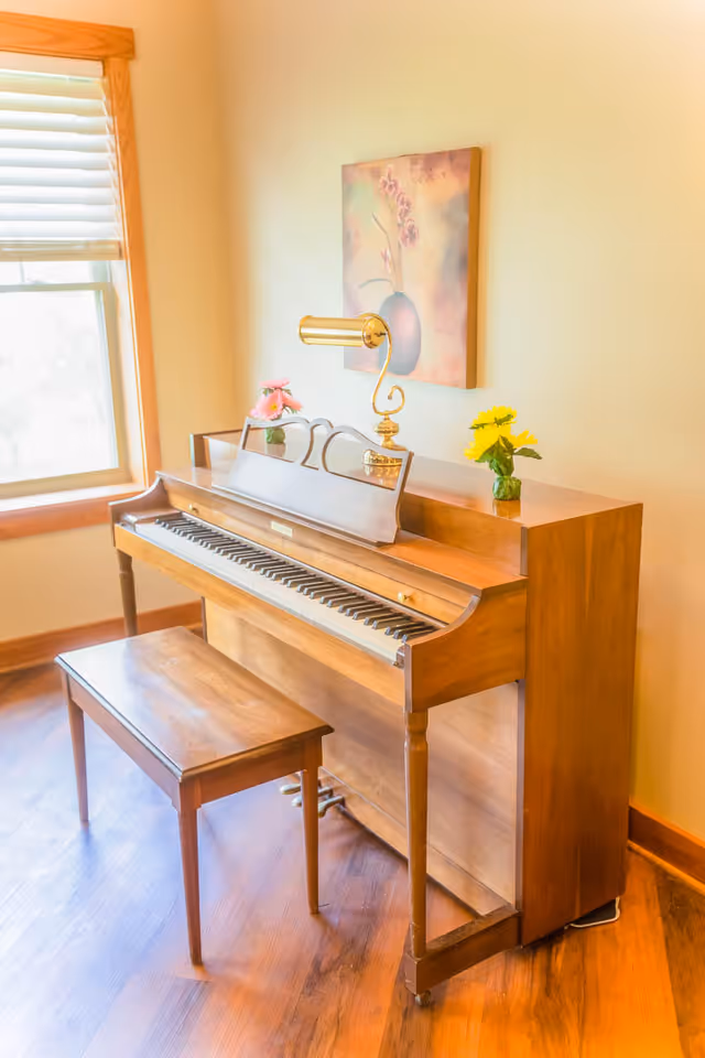 A wooden upright piano with a matching bench in a room with wooden flooring and a window with blinds. On top of the piano are a brass lamp and two small vases with flowers. A framed floral painting hangs on the wall above the piano.