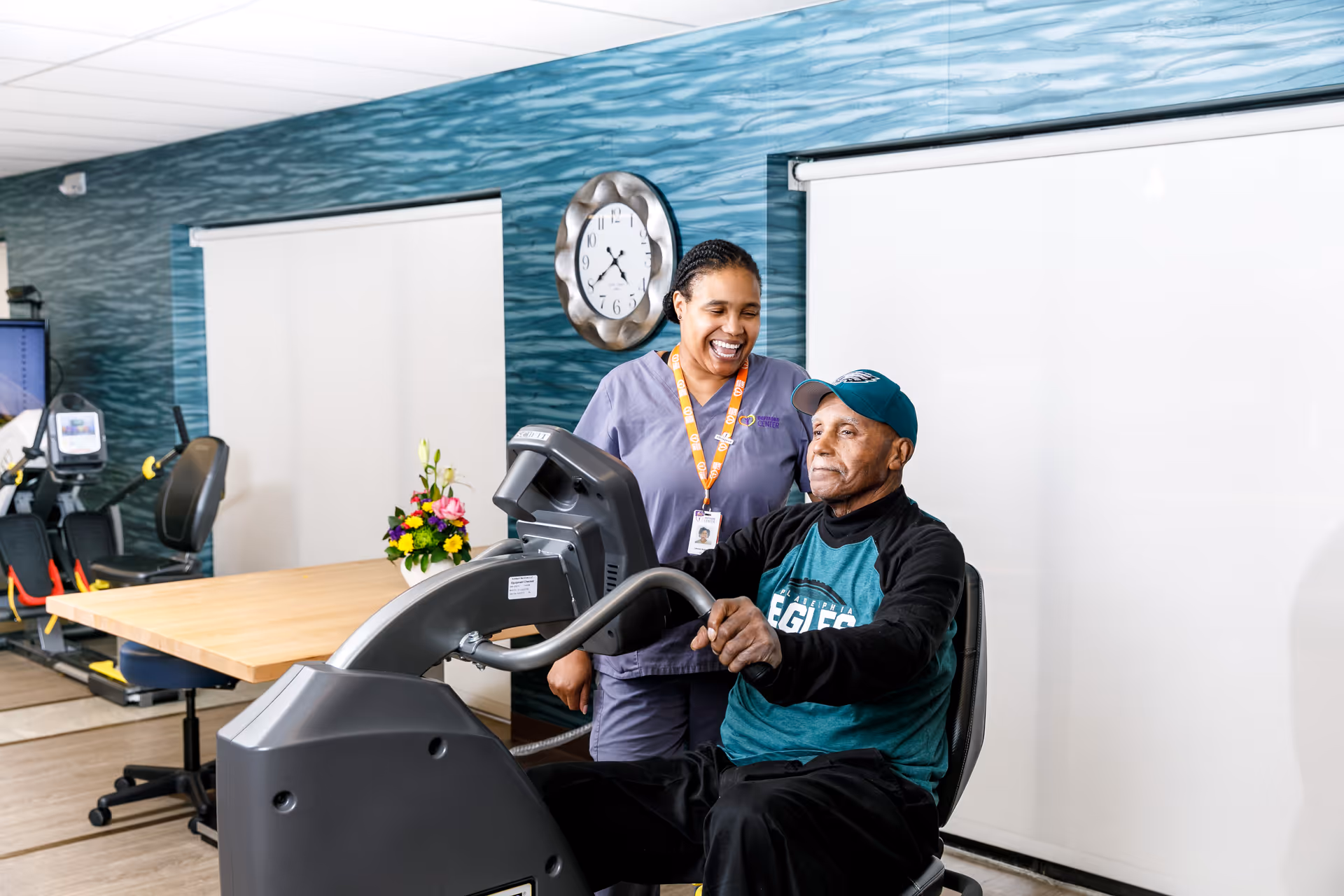 An elderly man wearing a green Eagles sweatshirt and cap is seated on a recumbent exercise bike while a smiling female caregiver in purple scrubs stands beside him, assisting. The room has a blue textured wall with a clock, a wooden table with a flower arrangement, and exercise equipment in the background.