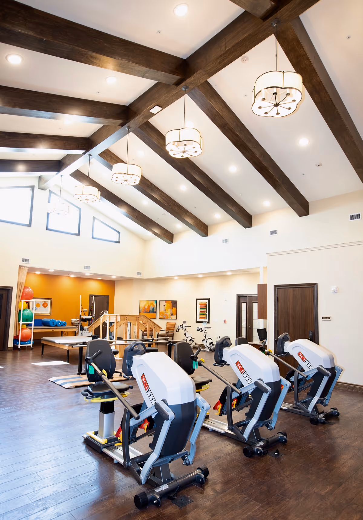 A spacious rehabilitation or physical therapy room with wooden beams on the ceiling and modern pendant lights. The room contains several exercise machines, including three seated rowing machines in the foreground, stationary bikes, and various therapy equipment like balance balls and parallel bars in the background. The walls are decorated with colorful framed artwork, and the floor is made of dark wood.